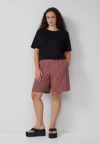 Schwarzes Kurzarmoberteil, lockerer Schnitt, kombiniert mit rosabraunen Shorts mit Taschen aus strukturiertem Stoff. Schwarze Plateausandalen.