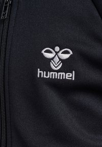 Veste noire zippée avec un logo blanc brodé présentant un design de abeille stylisé, accompagné du mot "hummel" en dessous. Tissu doux et texturé.