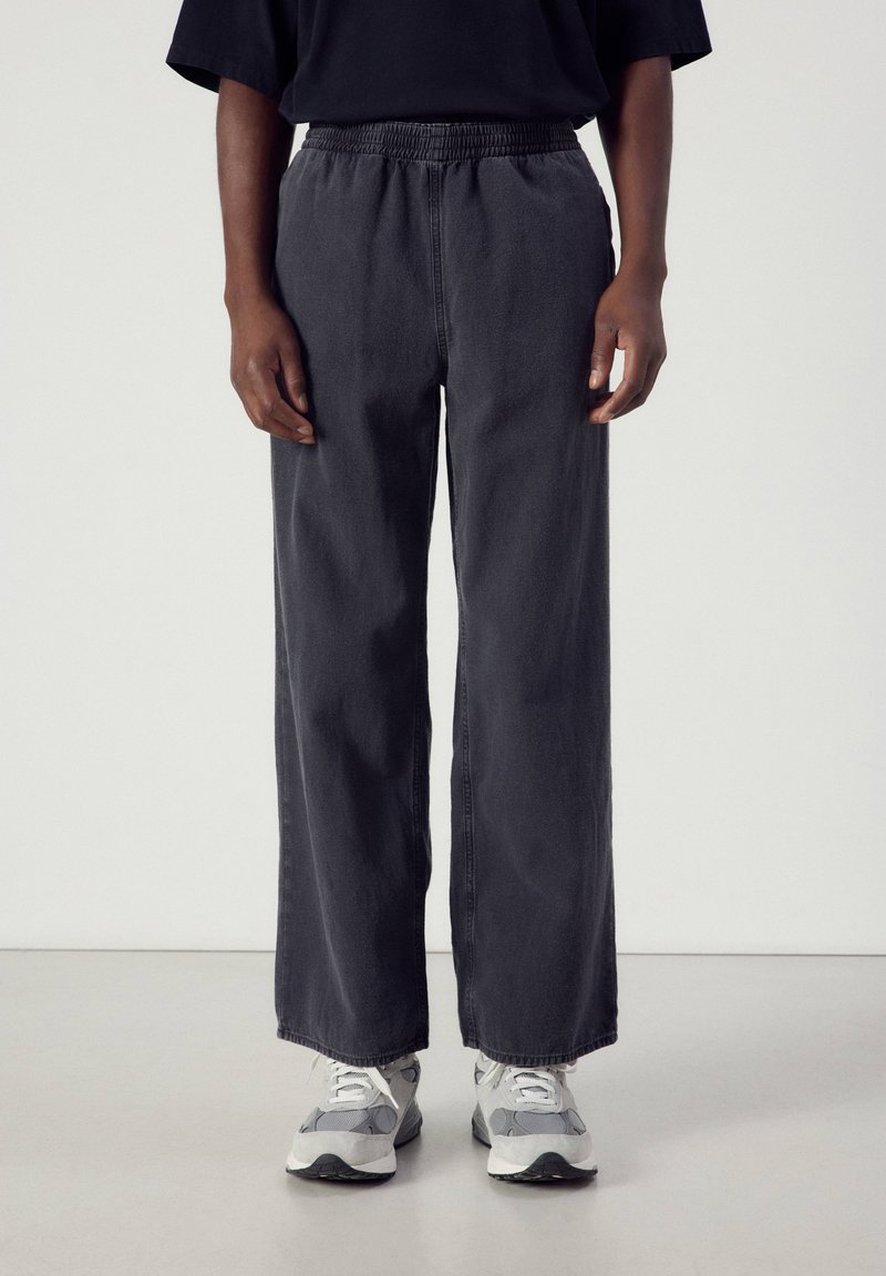 Personne portant un pantalon ample gris foncé à taille élastique et des baskets gris et blanches sur un fond clair uni.