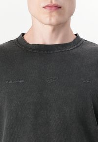 T-shirt en coton gris foncé avec un col rond côtelé, présentant des détails de logo subtils et la date d'établissement imprimée sur le devant. Texture douce.