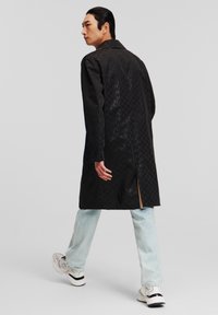 Abrigo negro largo con textura estampada, con cuello y abertura lateral. Llevado sobre unos vaqueros de color azul claro y zapatillas blancas.