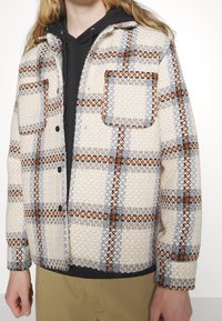 Skjorta-jacka i ullblandning i ett krämfärgat, grått och orange rutmönster. Har två bröstfickor, knappstängning och en kontrasterande svart hoodie under.
