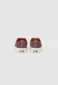 Low-top sneakers in a muted purple suede with orange accents and white rubber sole, featuring the "Converse All Star" logo on the heel.
