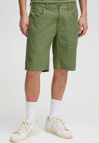 Žalios spalvos lina pagaminti šortai laisvo silueto, sukišti įkyrios kišenės priekyje ir užsegami sagomis. Dėvimi su baltais kojinėmis ir kreminės spalvos sportbačiais.