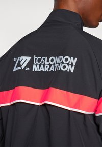 Black sports jacket featuring a red horizontal stripe and reflective text reading "TCS LONDON MARATHON 2025" on the back. Smooth fabric.