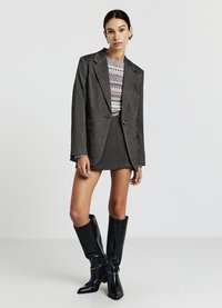 Blazer gris à revers, associé à une mini-jupe grise assortie. Le mannequin porte des bottes noires montantes avec une finition brillante et des embellissements.