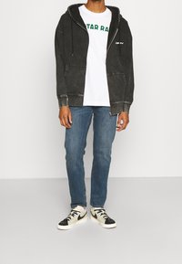 Felpa con cappuccio in denim nero con dettagli grigi, T-shirt bianca con scritta verde, jeans blu e sneakers nere e bianche, in piedi su uno sfondo neutro.