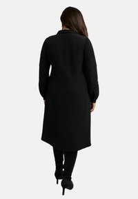 Černý dlouhý kabát s límcem, zdobený vyšívanými detaily na rukávech a s rovnou siluetou, v kombinaci s těsnými černými kalhotami.