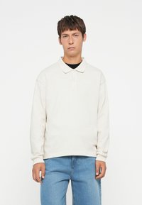Polo shirt bej deschis cu mânecă lungă, cu o croială confortabilă, guler și o clapetă cu două butoane. Asociat cu blugi din denim albastru deschis.