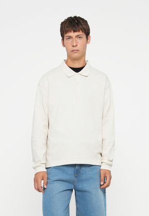 Light beige long-sleeve polo shirt with a relaxed fit, collared neck, and two-button placket. Paired with light blue denim jeans.
