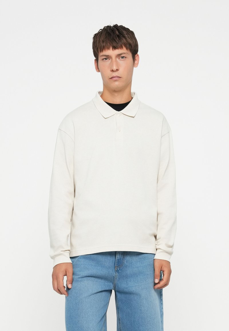 Polo shirt bej deschis cu mânecă lungă, cu o croială confortabilă, guler și o clapetă cu două butoane. Asociat cu blugi din denim albastru deschis.