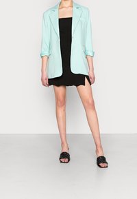 Mint green blazer with rolled sleeves, worn over a black mini dress. Black woven slide sandals complete the outfit.