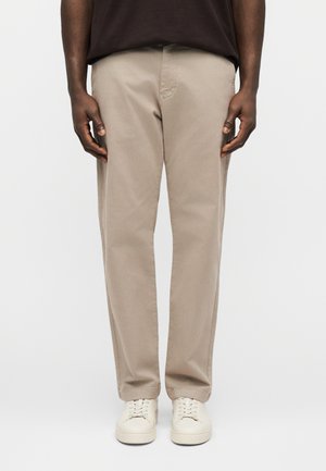 Mężczyzna w beżowych prostych spodniach, białych tenisówkach i ciemnobrązowej koszuli, stojący z rękami swobodnie opuszczonymi wzdłuż ciała na jasnym tle.