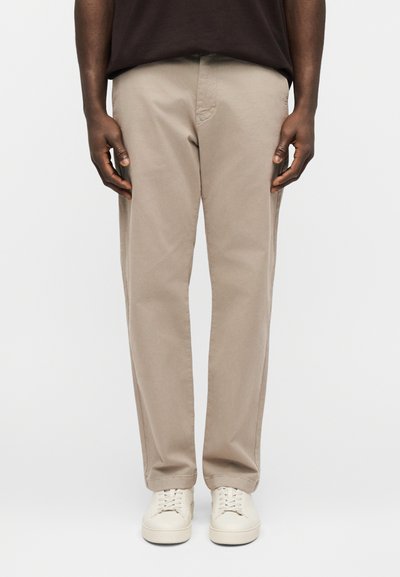 Homme portant un pantalon beige droit, des baskets blanches et une chemise marron foncé, debout avec les mains détendues le long du corps sur un fond clair.