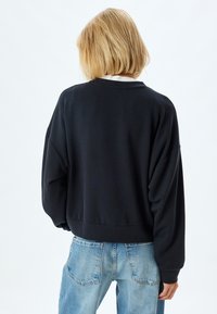 Schwarzes Sweatshirt mit lockerem Schnitt, überschnittenen Schultern und geripptem Saum. Kombiniert mit hellblauen Jeans, die einen lässigen Stil zeigen.