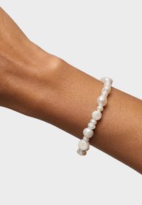 Nadgarstek o średnim brązowym kolorze skóry noszący białą nieregularną bransoletkę z pereł słodkowodnych na jasnym, jednolitym tle.