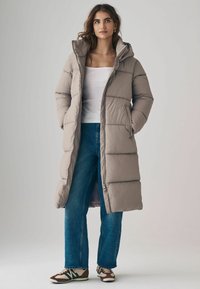 Frau in einem langen beigen Puffermantel mit Kapuze, weißem ärmellosem Oberteil, blauen Jeans und braunen sowie creamfarbenen Sneakers, die mit den Händen in den Taschen steht.
