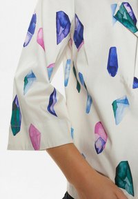 Blusa bianca con un motivo a cristallo acquerellato in viola, rosa, blu e verde. Maniche a tre quarti con taglio dritto.