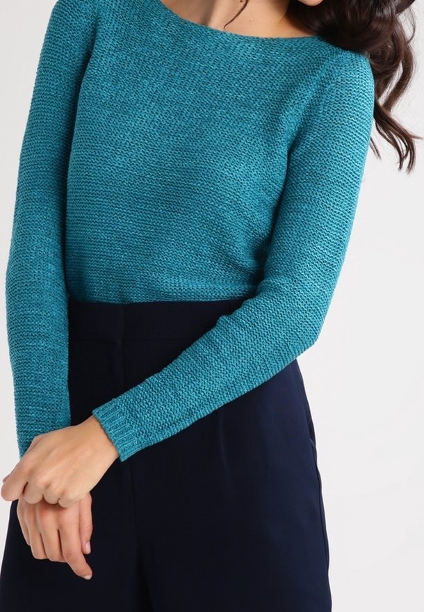 Femme portant un pull en maille texturée bleu sarcelle à manches longues, associé à un pantalon taille haute bleu marine foncé, debout les mains jointes.