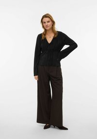 Svart plissért bluse med V-hals og knyting, kombinert med brune bukser med vide ben. Antrekket har lange ermer og teksturert stoff.