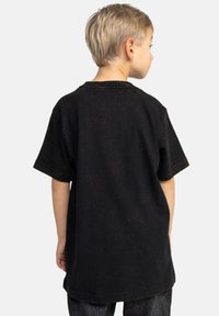 Černé bavlněné tričko s uvolněným střihem, krátkým rukávem a viditelnými švy, které má na sobě dítě obrácené zády k fotoaparátu.