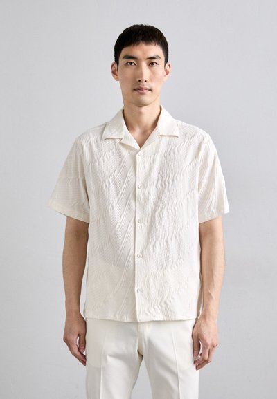 Chemise à manches courtes, boutonnée, en tissu texturé blanc cassé avec un motif ondulé et un col classique. Associée à un pantalon crème.