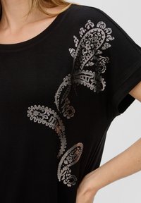 Czarny t-shirt z krótkim rękawem, ozdobiony metalicznym srebrnym wzorem na lewym ramieniu. Wykonany z miękkiego, lekkiego materiału.