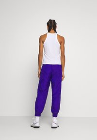 Bílý tílko s úzkými zády spojené s jasně fialovými, volně střiženými tepláky. Oblečení má elastické manžety a hladký povrch.