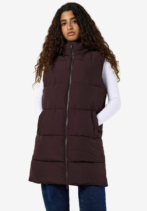Ung kvinde med langt krøllet hår iført en ærmeløs mørkebrun polstret vest over en hvid bluse med lange ærmer og blå jeans.