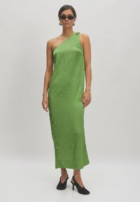 Asymmetrisches grünes Kleid mit einer gekräuselten Textur, aus leichtem Stoff gefertigt. Getragen mit schwarzen Sandalen mit offenen Zehen und Sonnenbrille.