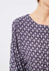 Mujer vestida con una blusa azul oscura con un patrón floral blanco y un escote redondo, mostrada desde el hombro hasta la mitad del pecho.