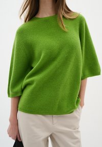 Green knit sweater with a relaxed fit, short sleeves, and a textured pattern. Paired with light beige trousers and a black handbag.
