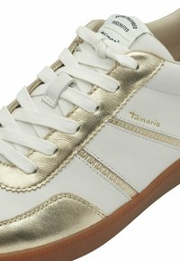 Baskets Tamaris blanches et dorées avec lacets blancs, accents dorés et semelle marron, affichant le nom de la marque sur le côté et la languette.