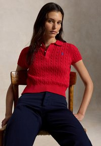 Žena s dlhými tmavými vlasmi, oblečená v červenom pánskom pulóvri s vrkočovým vzorom a námorníckych nohaviciach, sediaca na drevenej stoličke pred jednoduchým pozadím.