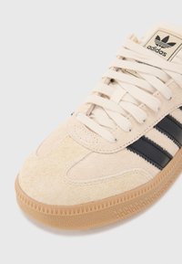 Beige suede sneaker with three black stripes, textured toe cap, and gum rubber sole. Features cream laces and logo on the tongue.