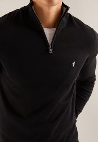 Černý svetr na zip s vysokým límcem, vyrobený z měkkého materiálu. Na hrudi má malé bílé logo ptáka.