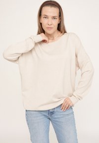 Beige übergroßer Pullover mit langen Ärmeln, Bootsausschnitt und gerippten Bündchen, kombiniert mit hellblauen Jeans, die einen entspannten Sitz schaffen.