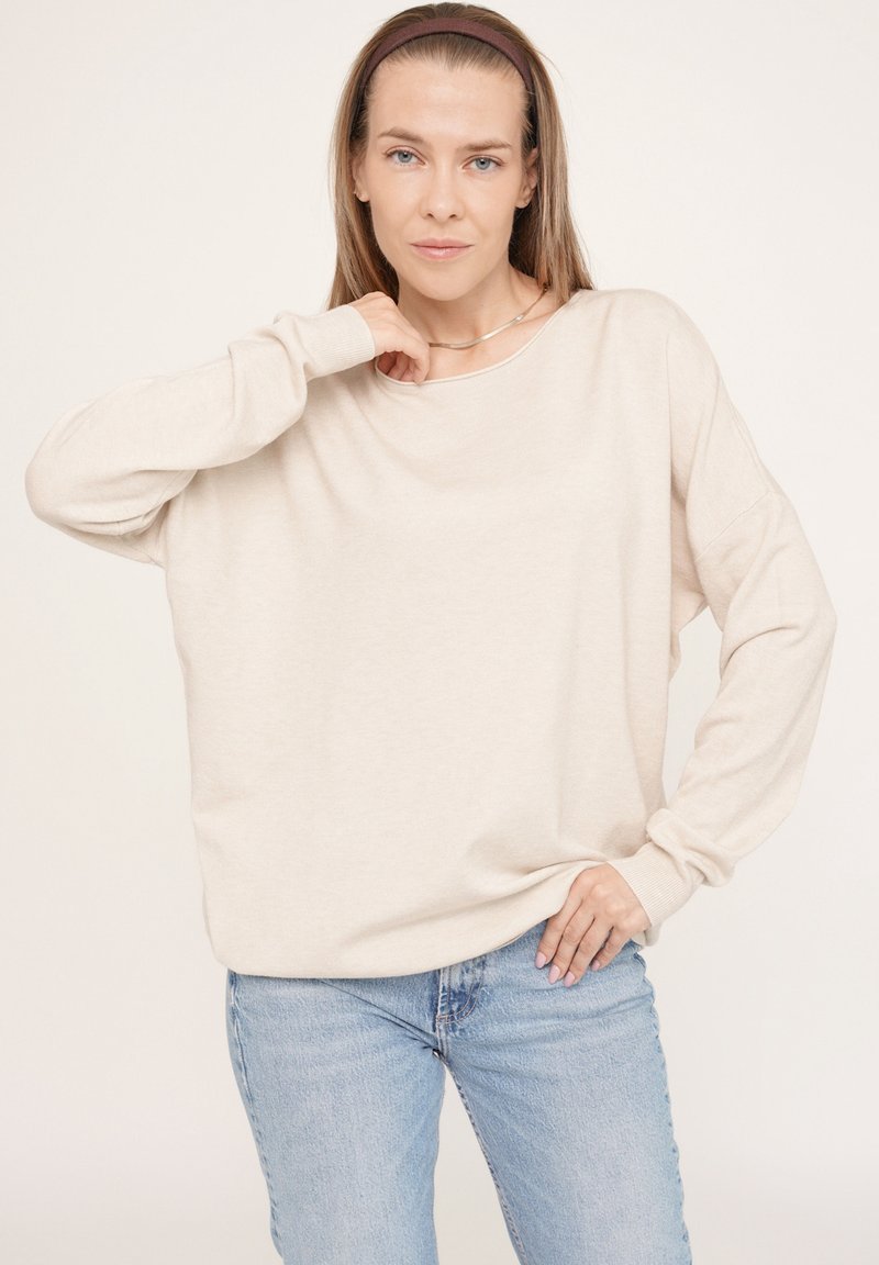 Beige übergroßer Pullover mit langen Ärmeln, Bootsausschnitt und gerippten Bündchen, kombiniert mit hellblauen Jeans, die einen entspannten Sitz schaffen.