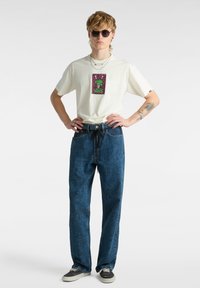 Weißes T-Shirt mit buntem Grafikdesign auf der Brust, kombiniert mit dunkelblauen Jeans mit schwarzen Mustern. Accessoires umfassen Sonnenbrillen und Halsketten.
