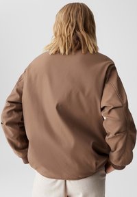 Bruine bomberjack met een gladde, matte textuur, met ribgebreide manchetten en een ontspannen pasvorm. Achteraanzicht toont een minimalistisch ontwerp en zachte stof.