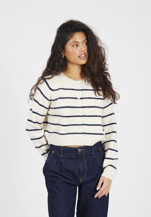 Vrouw met lang krullend haar, gekleed in een crèmekleurige trui met marineblauwe strepen en donkergame hoge taillebroeken, kijkend naar haar linkerkant.