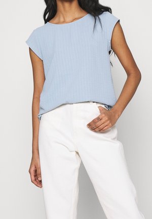 Woman wearing light blue short-sleeve textured blouse tucked into high-waisted white pants, hand resting in front pocket, neutral background.