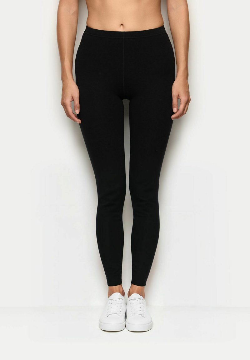 Leggings neri con un design aderente, realizzati in tessuto elasticizzato. Presentano una vita alta e una texture liscia, abbinati a sneakers bianche.