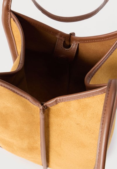 Καφέ σουέντ τσάντα tote με δερμάτινη επένδυση και λουριά, εμφανίζοντας ανοιχτό επάνω μέρος και ευρύχωρο εσωτερικό σε λευκό φόντο.