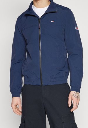 Mand iført en marineblå lynlåstrøje med logopatche på bryst og ærme, kombineret med sorte cargobukser.