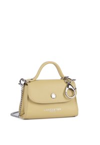 Petit sac à main Lancaster Paris beige avec une anse supérieure, une fermeture argentée et une bandoulière chaîne attachée sur fond blanc.