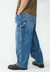 Jean oversized en denim bleu avec une coupe décontractée, comportant une poche latérale, des coutures contrastées et une finition usée, accompagné de mocassins marron.