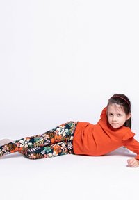 Czerwona koszula z długim rękawem i falbankami na ramionach, w połączeniu z kolorowymi legginsami w kwiatowe wzory w kolorach pomarańczowym, różowym i zielonym.