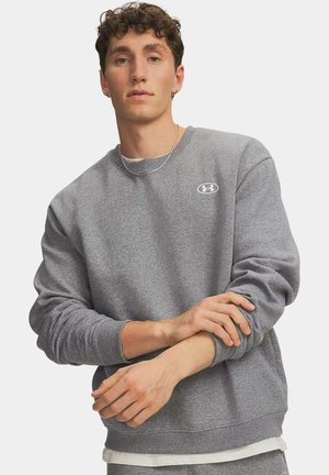 Jonge man met krullend haar, gekleed in een grijze Under Armour trui, die zijn mouw aanpast tegen een effen achtergrond.
