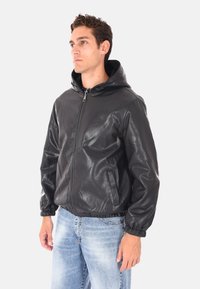 Uomo che indossa una giacca di pelle nera con zip e cappuccio, insieme a jeans azzurri chiari, in piedi e rivolto verso sinistra su uno sfondo neutro.